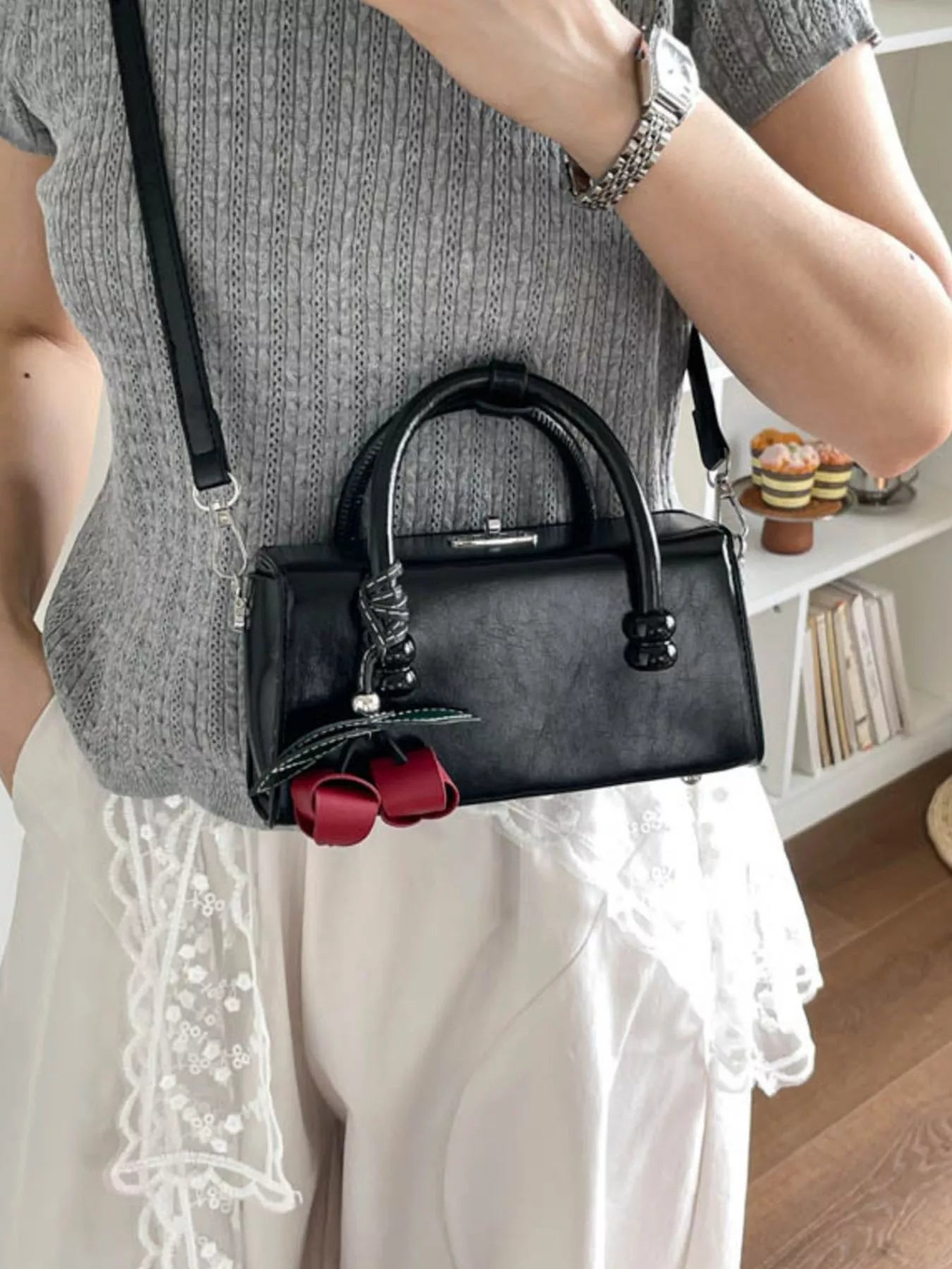 Black top-handle crossbody bag with shiny hardware and red bow charm.