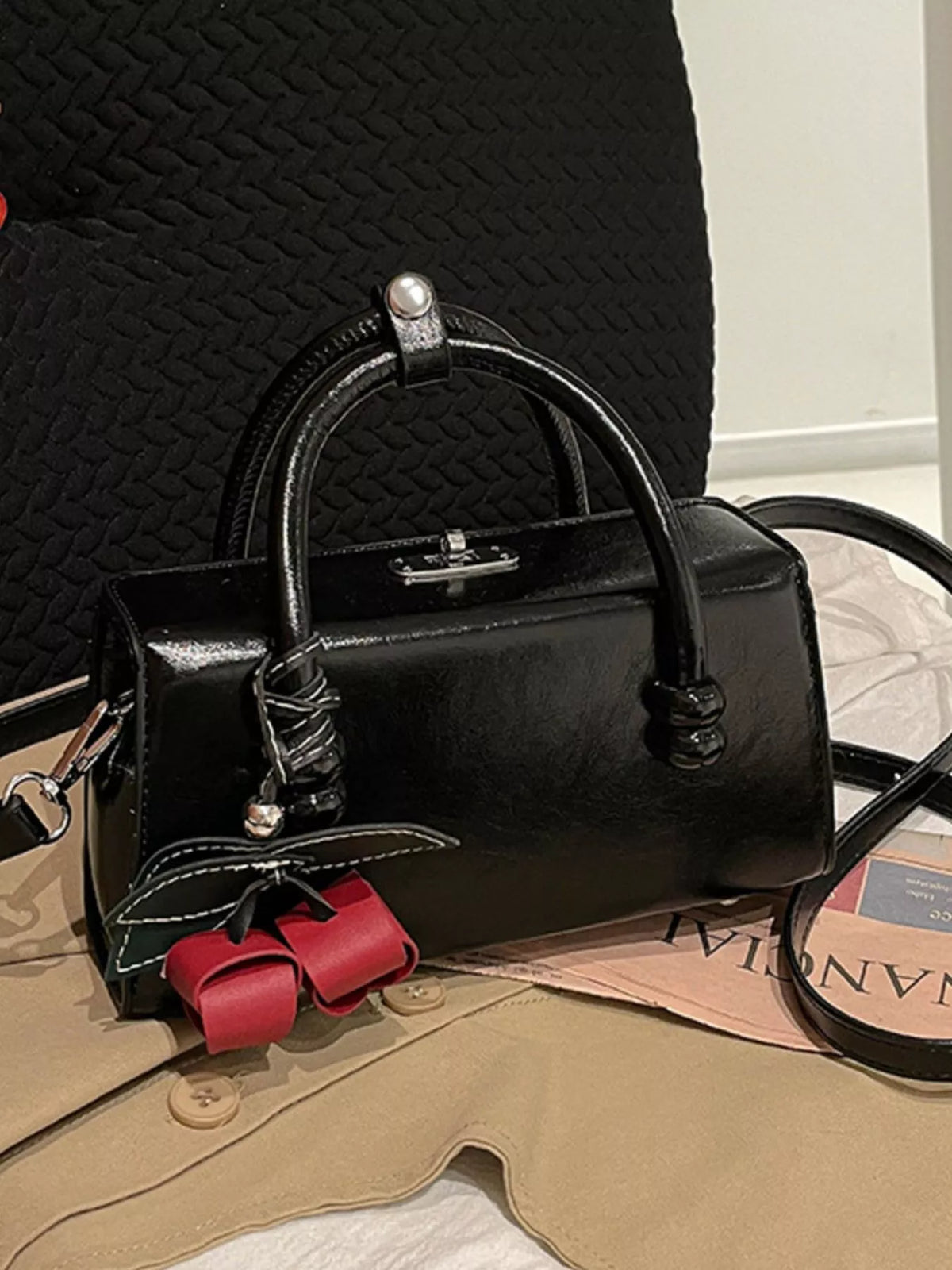 Black top-handle crossbody bag with red bow charm and textured front detail.