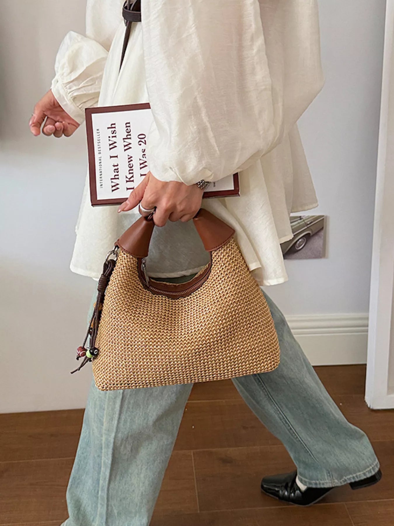 Tan woven straw crossbody bag with brown leather handles and dark beaded tassel on the side